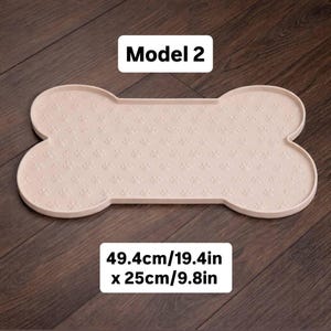 May include: A light pink, bone-shaped pet food mat with a paw print pattern. The mat is 19.4 inches long and 9.8 inches wide. The text "Model 2" is displayed on the mat.