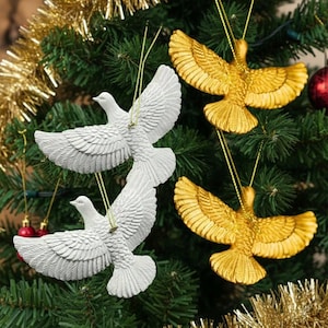 May include: Christmas tree ornaments featuring white and gold bird designs. The white birds are in flight with detailed wing feathers. The gold birds have outstretched wings. The ornaments hang from gold strings against a backdrop of green pine needles and gold tinsel.
