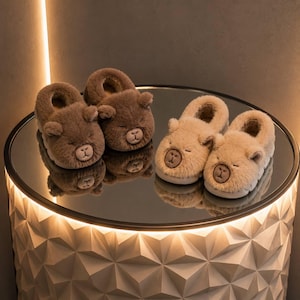 Puede incluir: Dos pares de pantuflas de felpa con forma de cara de oso, un par en marrón oscuro y otro en beige claro. Cada pantufla presenta orejas redondeadas, ojos cerrados bordados y un detalle de nariz cosido en la superficie exterior suave y peluda.