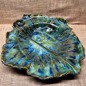 May include: A ceramic leaf-shaped serving dish with a glossy finish. The dish features a blend of blue, green, and gold colours, with intricate veining details. The edges are slightly ruffled, and the dish has a loop on one side.