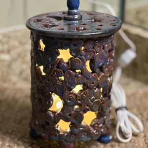 Puede incluir: Un calentador eléctrico de cerámica con un esmalte azul y marrón. El calentador tiene un diseño decorativo de remolinos y tiene una luz en el interior. El calentador está sobre una superficie tejida.