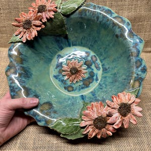 Puede incluir: Un cuenco de cerámica hecho a mano con un esmalte turquesa y un borde ondulado. El cuenco está adornado con girasoles tridimensionales de color melocotón y hojas verdes. El cuenco está sostenido por una mano.