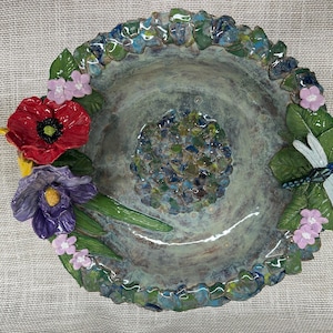 Puede incluir: Un bebedero para pájaros de cerámica hecho a mano con una cuenca central y un borde decorativo. El borde está adornado con hojas esculpidas, flores y una libélula. El bebedero para pájaros presenta una amapola roja, un iris morado y flores rosas. Diámetro de unos 30 cm.