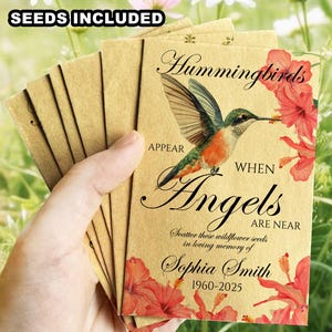 May include: A stack of seed packets with a hummingbird illustration and text. The packets are tan with red floral accents and the words "Hummingbirds Appear When Angels Are Near". The text "Seeds Included" is at the top.