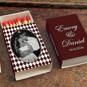 May include: A burgundy matchbox with a black and white checkered pattern and a photo of a couple kissing. The text "Emery & Daniel" and the date "06.16.2026" are on the matchbox. The match heads are red.