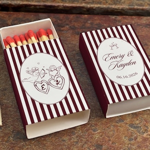 May include: Two matchboxes with a burgundy and white striped design. One box is open, revealing red-tipped matches. The boxes feature a white oval with a cupid illustration and the names "Emery & Kayden" with a date. Two loose matches are visible.