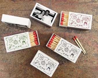 May include: White matchboxes with red-tipped matches. Some boxes feature a black and white photo, while others have a decorative design with a disco ball, champagne glasses, and the date 07.18.26. The matches are arranged inside the boxes.