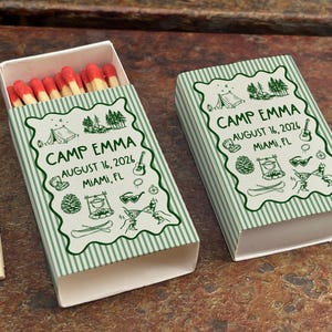 Op de afbeelding: Twee lucifersdoosjes met een groen en wit gestreept ontwerp. De dozen zijn voorzien van de tekst "CAMP EMMA AUGUST 16, 2026 MIAMI, FL" in een decoratief lettertype. E&eacute;n doosje is open, met rode lucifers.