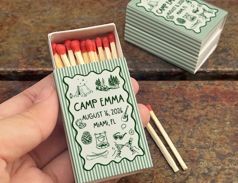 Op de afbeelding: Een luciferdoosje met een groen en wit gestreept ontwerp, met de tekst "CAMP EMMA AUGUST 16, 2026 MIAMI, FL" en illustraties met kampeerthema. De doos is open en onthult lucifers met rode uiteinden.