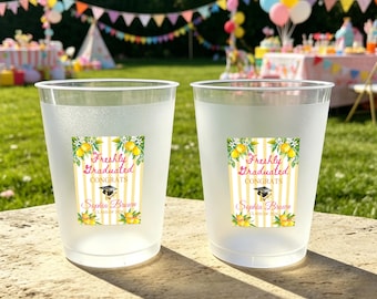 Op de afbeelding: Twee matte plastic bekers met labels met een diploma-uitreikingsthema. De labels bevatten de tekst "Freshly Graduated Congrats" met een illustratie van een baret, citroen- en bloemaccenten en een naam. De bekers staan op een grasveld.