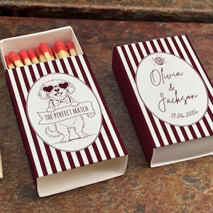 May include: A maroon and white striped matchbox with a cartoon dog wearing heart-shaped sunglasses and the text "THE PERFECT MATCH". The closed matchbox has the names "Olivia & Jackson" and the date "11.06.2026".