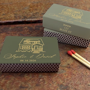 May include: Olive green matchboxes with gold lettering and a house illustration. The text on the boxes reads "Sophie & David" and "06.16.2026", with the address "123 Main St" on one box. Two matches with red tips are visible.