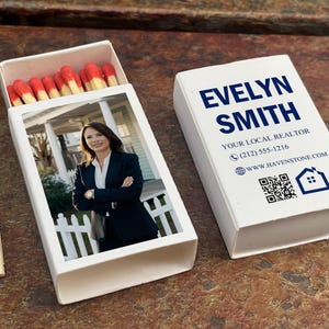May include: White matchbox with the name "EVELYN SMITH" in blue text. The box also includes contact information and a QR code. Inside, matches with red tips are visible. The open matchbox displays a photo of a woman in a suit.