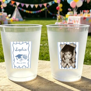 May include: Two frosted plastic cups with graduation-themed stickers. One cup has a sticker with the name "Sophia", a graduation cap, and the text "Class of 2026". The other cup features a photo of a child in a graduation cap.