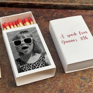 May include: White matchbox with a black and white photo of a person wearing sunglasses. The matchbox contains red-tipped matches. The closed matchbox has the text "A spark from Gianna's 30th" in red script.