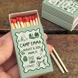 Op de afbeelding: Een luciferdoosje met een groen en wit gestreept ontwerp, met de tekst "CAMP EMMA AUGUST 16, 2026 MIAMI, FL" en illustraties met kampeerthema. De doos is open en onthult lucifers met rode uiteinden.