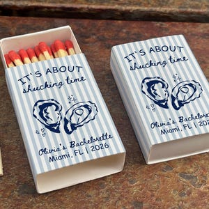 May include: Two rectangular matchboxes with light blue and white vertical stripes. The text reads IT'S ABOUT shucking time, Olivia's Bachelorette, Miami, FL | 2026, alongside illustrations of two oysters. One box is open, revealing red-tipped matches.
