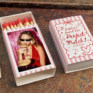 May include: A matchbox with a pink and white striped design, featuring a photo of a person holding a cocktail. The box is open, revealing red-tipped matches. The closed box has text that reads "Sophia's found her Perfect Match! 11.05.2026".