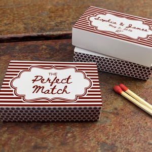 May include: Three rectangular matchboxes with red and white striped designs. One box reads "The Perfect Match" in a decorative font. Another box has the names "Sophia & James" and a date. Two matches with red tips are next to the boxes.