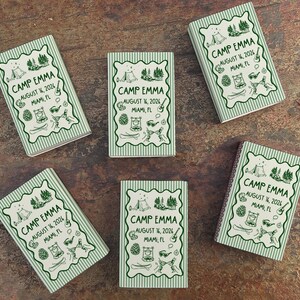 Op de afbeelding: Vijf kleine, rechthoekige notitieboekjes met een groen-witte gestreepte rand. Elk notitieboekje bevat de tekst "CAMP EMMA AUGUST 14, 2026 MIAMI, FL" in een decoratief kader met kampeerillustraties.