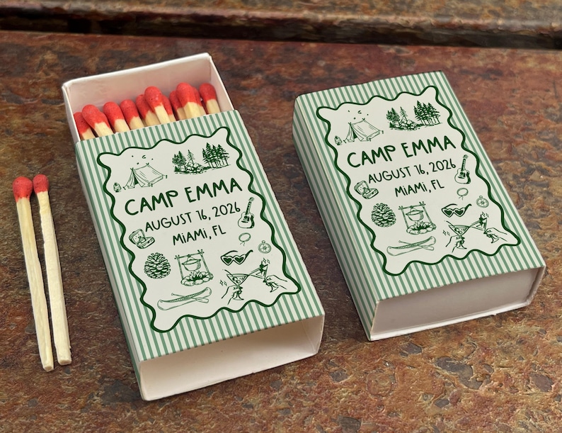 Op de afbeelding: Twee lucifersdoosjes met een groen en wit gestreept ontwerp. De dozen zijn voorzien van de tekst "CAMP EMMA AUGUST 16, 2026 MIAMI, FL" in een decoratief lettertype. E&eacute;n doosje is open en toont lucifers met rode uiteinden. Het andere doosje is gesloten.