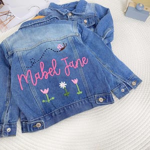 May include: Two blue denim jackets. The front jacket features the name "Mabel Jane" embroidered in pink, with a bee and floral designs. The second jacket is visible behind. Both jackets have button fastenings.