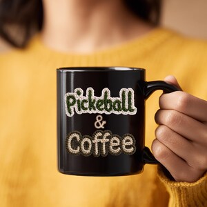 May include: Black coffee mug with the words "Pickeball & Coffee" in a crocheted style. The text is in green and brown colors. The mug is held by a person wearing a yellow sweater.