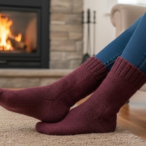 Pode incluir: Meias de malha bordô com punhos canelados. As meias grossas são de cor vermelho escuro. A pessoa está usando jeans azuis e sentada perto de uma lareira. As meias estão sobre um tapete bege.