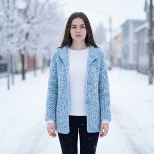 Könnte beinhalten: Hellblauer Strickcardigan mit Kragen und zwei Knöpfen. Der Cardigan wird über einem weißen Langarmshirt und einer schwarzen Hose getragen. Der Hintergrund zeigt eine verschneite Straßenszene.