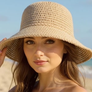 May include: A beige woven sun hat with a wide brim. The hat is worn outdoors on a sunny day. The hat is made of a light-colored, textured material, and the brim provides shade. The background includes a beach and ocean.