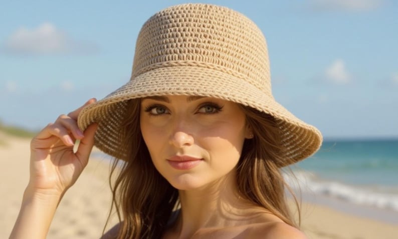 May include: A beige woven sun hat with a wide brim. The hat is worn outdoors on a sunny day. The hat is made of a light-colored, textured material, and the brim provides shade. The background includes a beach and ocean.