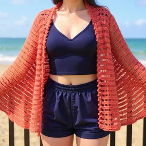 May include: A coral-coloured, open-front crochet cardigan with a scalloped hem. The cardigan has a textured, open weave design and is adorned with small, round embellishments along the edges. The model is wearing a navy blue top and shorts.