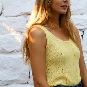 May include: A light yellow knitted tank top with a scoop neckline. The sleeveless top has a textured knit pattern with vertical lines of small holes. The garment is made of a soft, lightweight material.