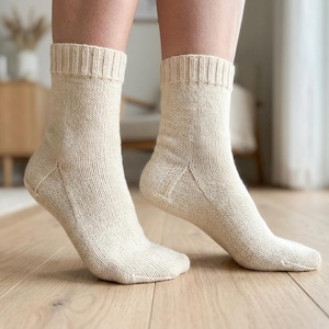 May include: A pair of cream-colored knitted socks. The socks are ribbed at the top and have a textured appearance. The socks are on a light-colored wooden floor.