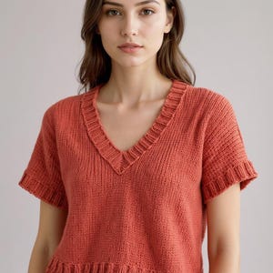 Puede incluir: Una blusa de punto color coral con cuello en V y mangas cortas. La blusa presenta un diseño acanalado a lo largo del escote, los puños de las mangas y el dobladillo. La tela parece ser un tejido de peso medio, adecuado para climas cálidos.