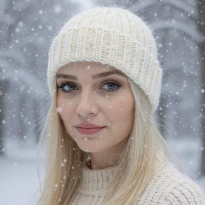 Puede incluir: Una mujer con un gorro y un suéter de punto color crema, con cabello largo y rubio, en un entorno nevado al aire libre. El gorro es acanalado y con vuelta. Los copos de nieve caen alrededor de la mujer.