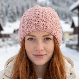 May include: A light pink, crocheted beanie hat. The hat is made of a soft, textured yarn and has a ribbed brim. The hat is worn by a person with long, red hair and freckles, with a snowy background.