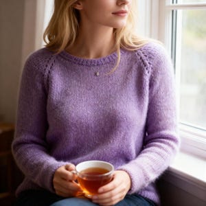 Peut inclure: Un pull violet clair avec un col rond et des manches raglan. Le pull est fait d'un tissu tricoté doux et texturé. Une tasse à thé en verre transparent remplie de thé est tenue dans les mains.