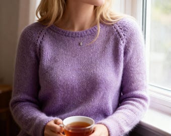 May include: A lilac-coloured knitted sweater with a round neckline and raglan sleeves. The sweater is made of a soft, textured yarn. A clear glass teacup filled with tea is held in the hands.