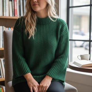 Puede incluir: Un suéter de punto acanalado verde oscuro con cuello alto. El suéter tiene mangas largas y lo lleva una persona sentada en una silla. Una estantería y una ventana son visibles en el fondo.