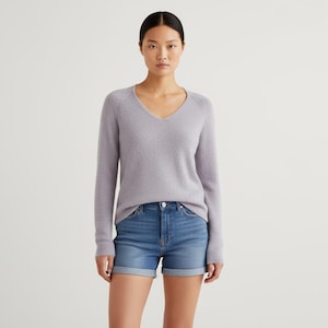 May include: A person wearing a pale purple V-neck jumper and blue denim shorts. The shorts have a turned-up hem. The background is plain white.