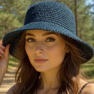 May include: A dark blue, woven sun hat with a wide brim. The hat is worn by a person in a natural outdoor setting. The hat's texture and shape are clearly visible, suggesting a casual, stylish accessory for sun protection.