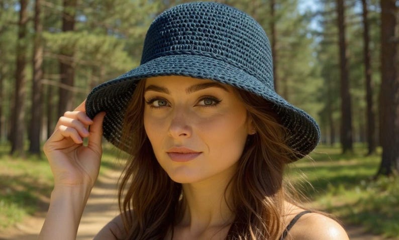 May include: A dark blue, woven sun hat with a wide brim. The hat is worn by a person in a natural outdoor setting. The hat's texture and shape are clearly visible, suggesting a casual, stylish accessory for sun protection.