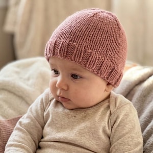 Może przedstawiać: Dzianinowa czapeczka dla dziecka w kolorze przydymionego różu. Czapeczka ma prążkowany brzeg i wzorzysty wzór dzianiny. Dziecko ma na sobie jasny pajacyk. Tło jest miękkie i rozmyte.