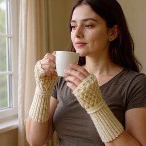 May include: Cream-colored, knitted fingerless gloves with a textured pattern on the hand and ribbed cuffs. The gloves are worn by a person holding a white mug near a window.