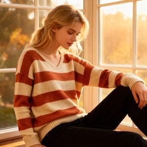 Könnte beinhalten: Ein V-Ausschnitt-Pullover mit breiten horizontalen Streifen in Rostorange und Creme. Der Pullover hat lange Ärmel und wird mit einer schwarzen Hose getragen. Die Person sitzt an einem Fenster.
