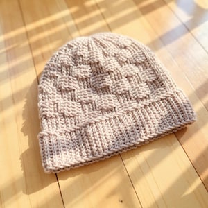 May include: A beige knitted beanie hat with a basketweave pattern. The hat has a ribbed brim and a textured crown. The hat is made of a soft, warm material, perfect for cold weather. The hat is shown on a wooden surface.