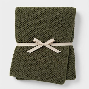 Può includere: Coperta di maglia verde oliva piegata con un motivo strutturato. La coperta è legata con un nastro e un fiocco beige. La coperta è realizzata in un materiale morbido e confortevole.