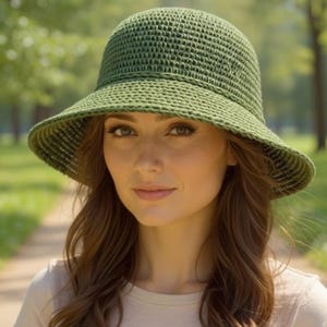 Puede incluir: Un sombrero de sol verde tejido a ganchillo con ala ancha. El sombrero está hecho de un material texturizado y tejido. El sombrero lo lleva una persona con el pelo largo y castaño. El fondo es una escena borrosa al aire libre con árboles.
