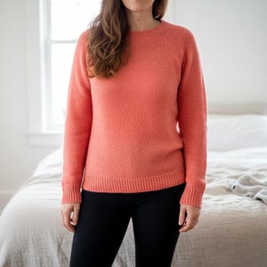 May include: A coral-colored sweater with a crew neck and long sleeves. The sweater has a textured knit pattern and is paired with black leggings. The background includes a bed with a white comforter and a window.